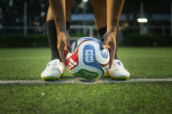A soccer player's hands lower a ball onto the grass pitch between his feet.