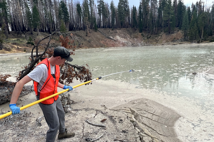 A researcher in an orange safety vest and blue gloves uses a long yellow pole to collect a water sample from the edge of Boiling Springs Lake, a pale gray-green body of water.