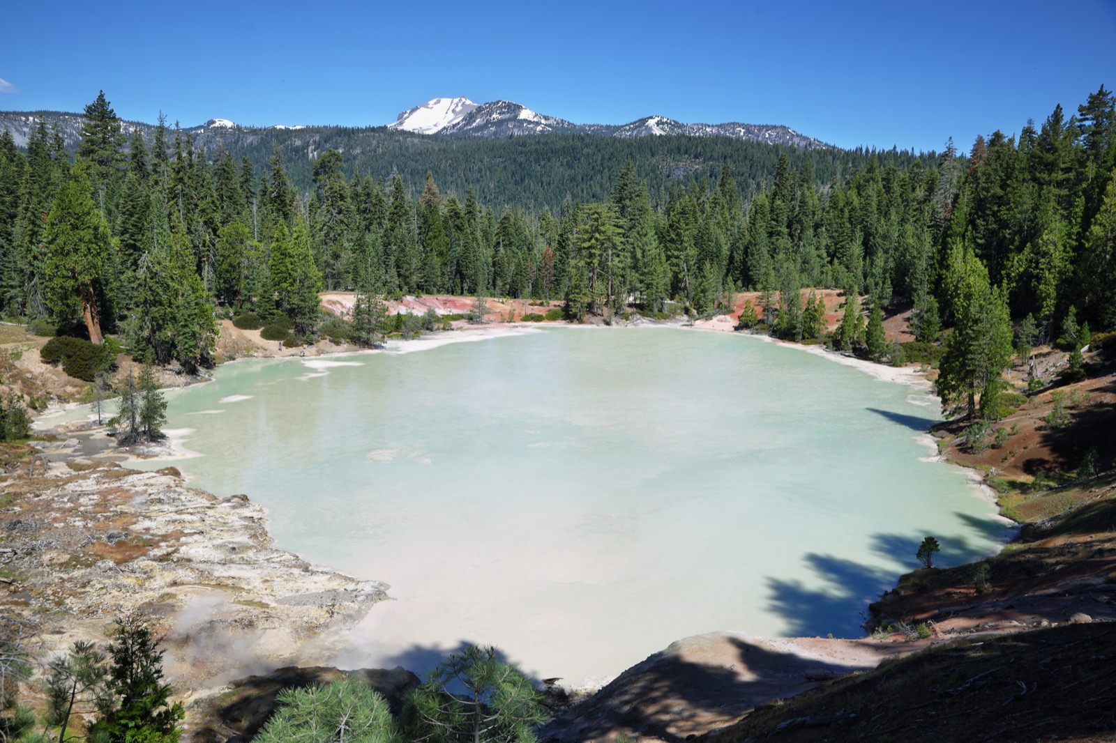 An aerial view of a large lake with pale green, milky water, surrounded by conifer forest and mineral deposits on a rocky shore.