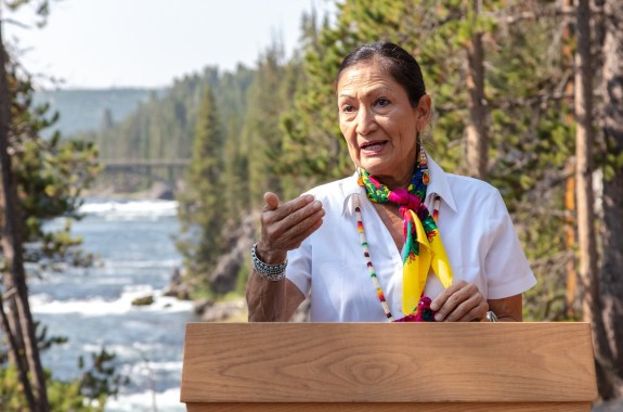 A woman in a white shirt gestures whike she speaks at a podium with a river and forest in the background.