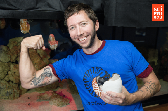 A man with brown hair and beard in a blue shirt holds a chambered nautilus while showing off his tattoo of the same animal.