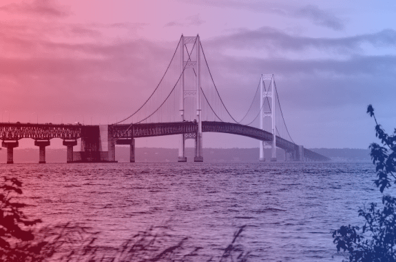 Scenic view of Mackinac Bridge spanning over the water on a cloudy day, framed by greenery with a pink and blue filter overtop the image.