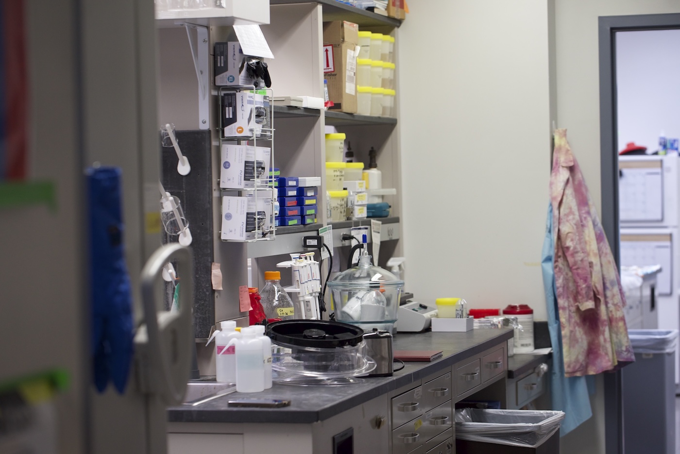 A view trough the door to a countertop along a lab wall, stacked with plastic containers, small boxes and other items. A pink lab coat hangs in the background.