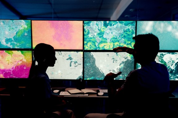 Two people sit in a dark room, illuminated by computer screens showing storms and weather patterns.