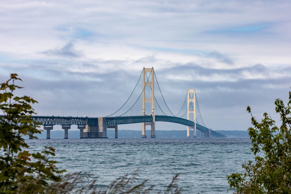 Scenic view of Mackinac Bridge spanning over the water on a cloudy day, framed by greenery.