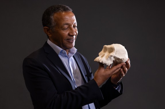 A man in a suit holds up a model of a an early human skull and looks at it, smiling