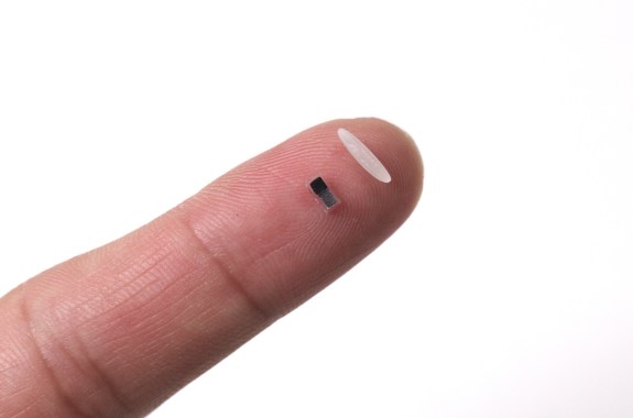 A tiny pacemaker on a finger next to a grain of rice.