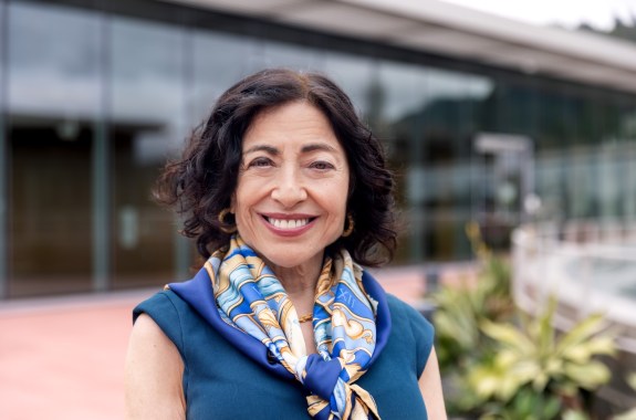 A woman with dark hair in a blue shirt and colorful scarf stands outside smiling at the camera.
