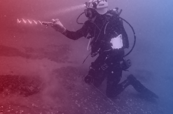 A diver kneels on the sandy bottom of a lake holding a tool with a corkscrew tip.