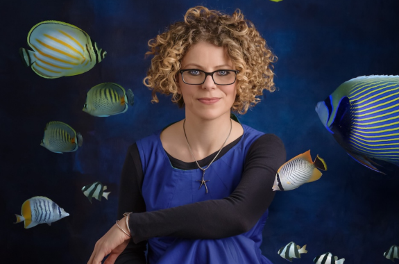 A women in a blue dress with curly, blonde hair and glasses sits amongst a group of different colored fish swimming around her in the air.