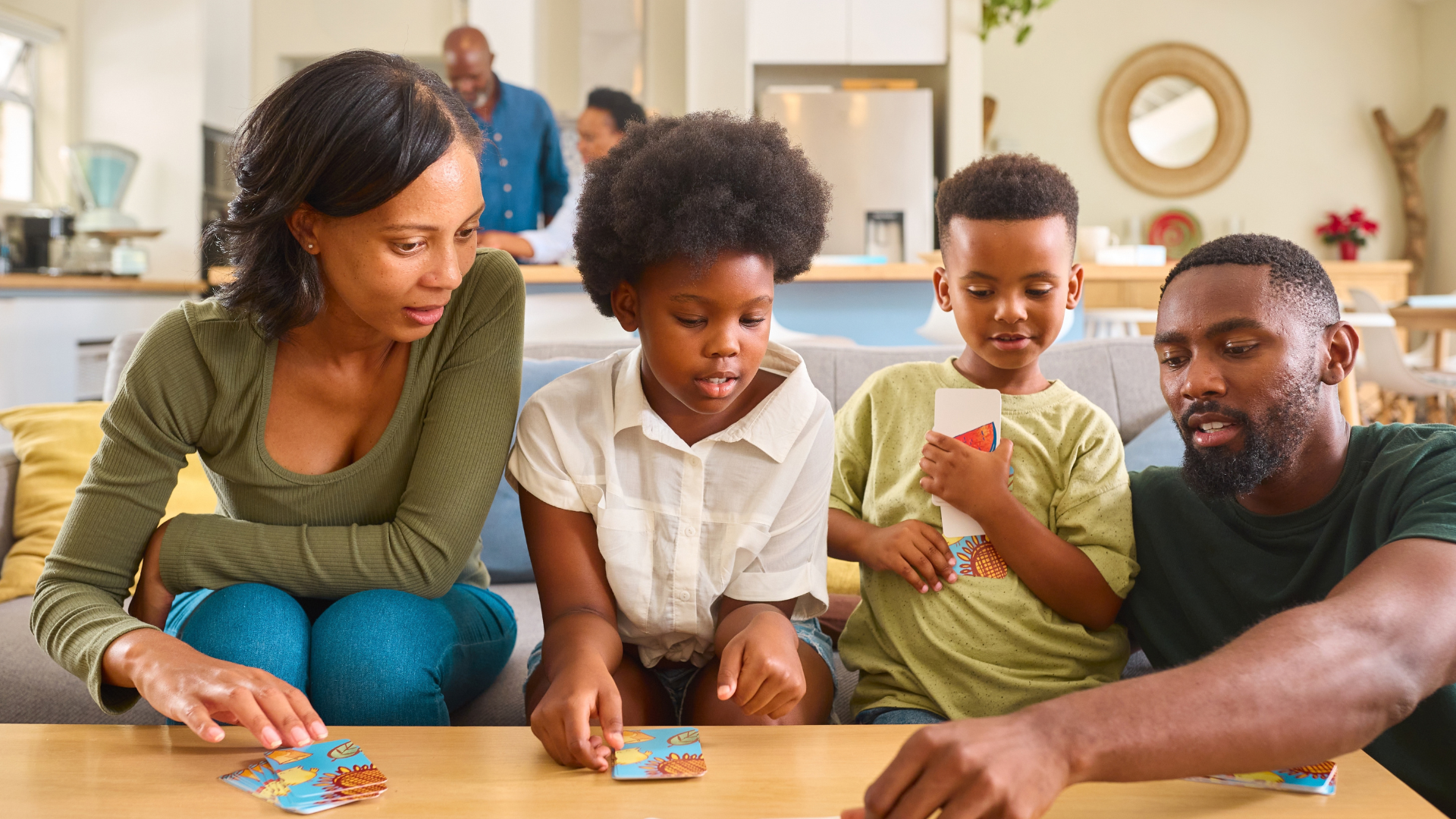 A family of four sits on a couch by a coffee table and plays a card game.