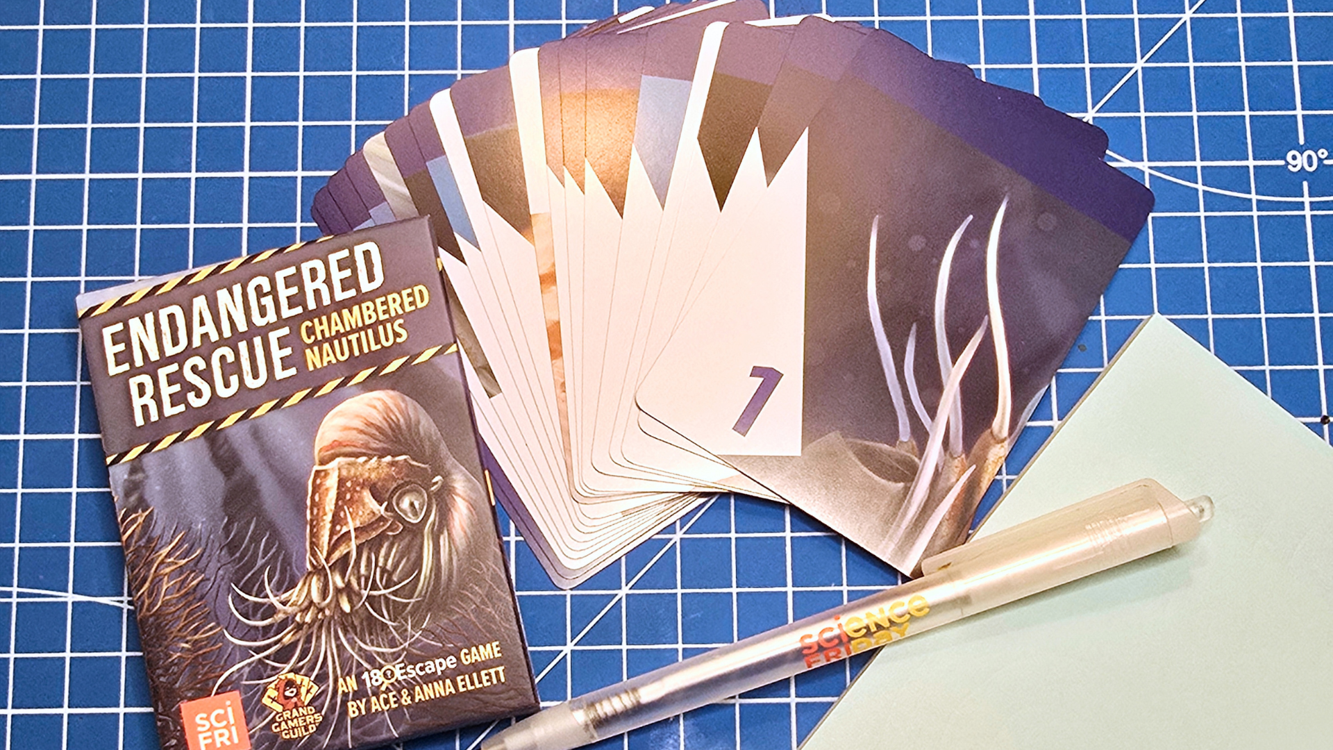 On a blue craft mat lies the box for the Endangered Rescue Chambered Nautilus game, the cards fanned out, a pen, and a pad of paper.