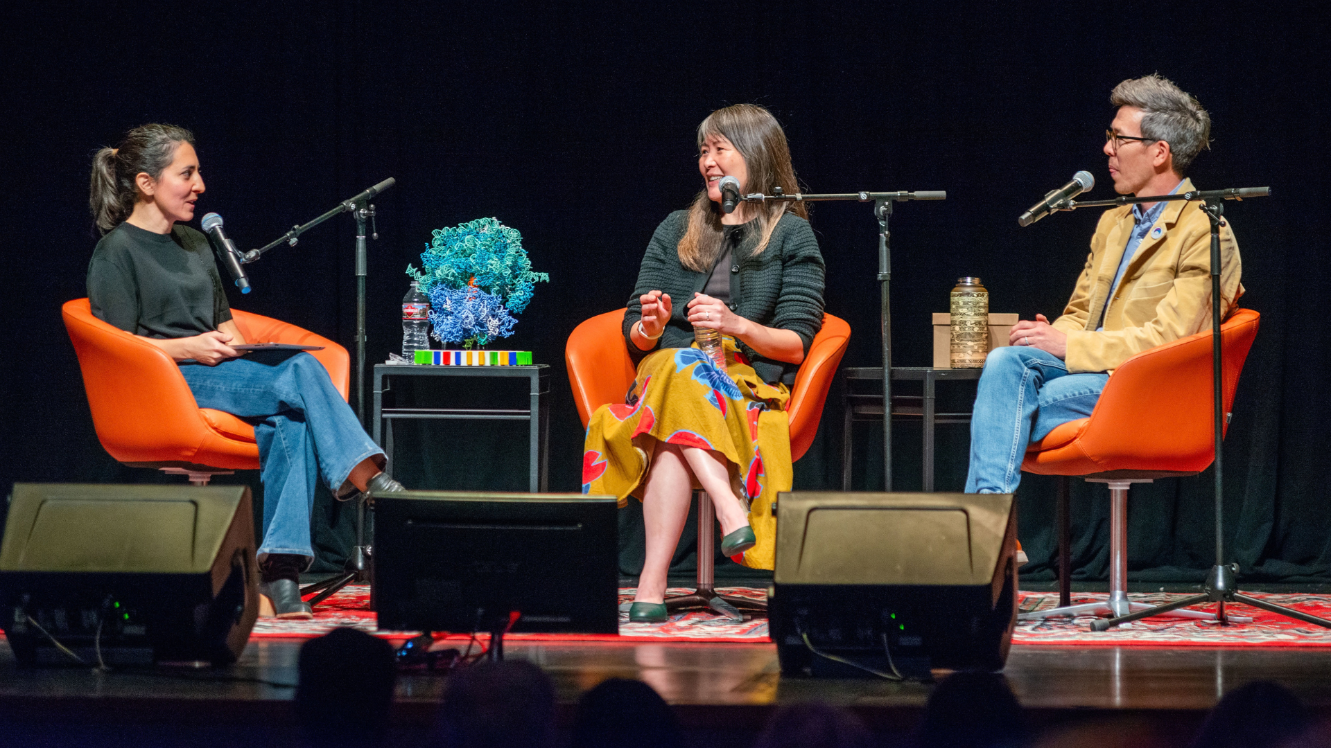 Three people talk to one another on a stage with microphones on stands in front of them.