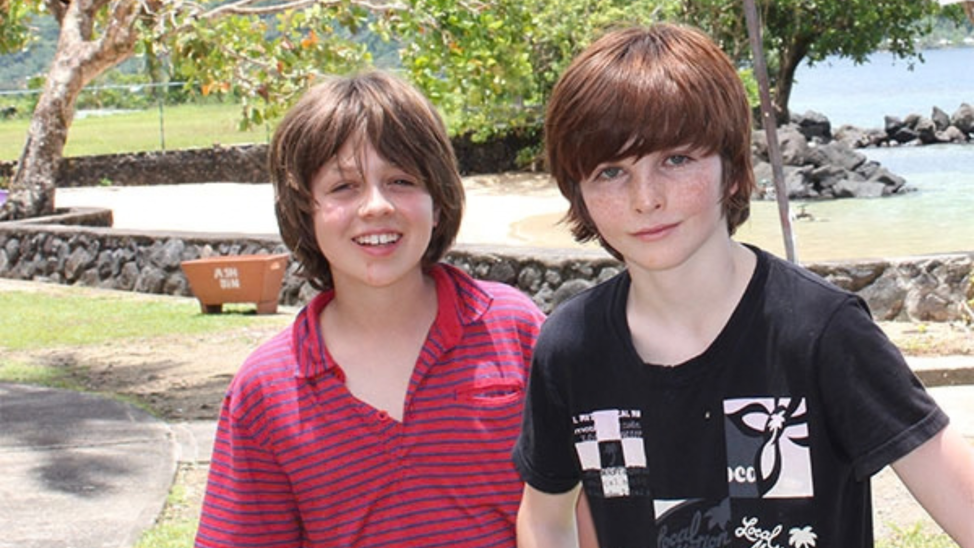 Two 11 year old boys stand outside with trees and the beach behind them.