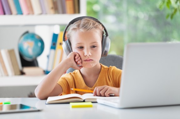 A young boy wearing headphones sits in front a bookcase looking at a laptop.