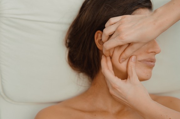 A brunette woman lays on a pillow while a woman uses her hands to massage her face as part of a lymphatic drainage procedure.