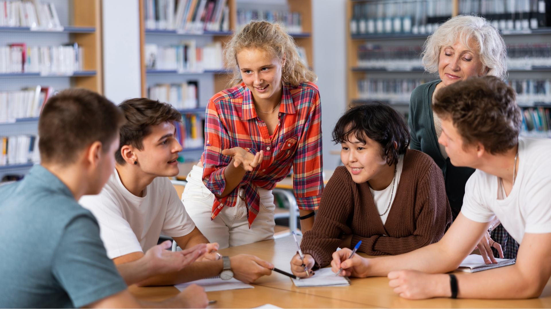 A group of five young teens sits around a table in the library, talking and writing notes. An adult teacher looks over their shoulders to observe what they are doing.