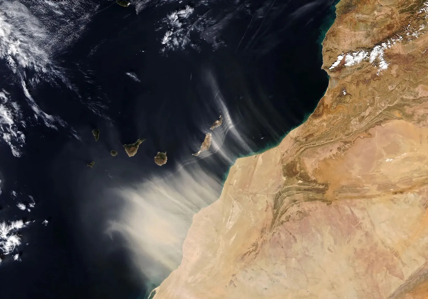 Aerial view showing a wash of pale dust sweeping from a brown landmass out over the dark ocean