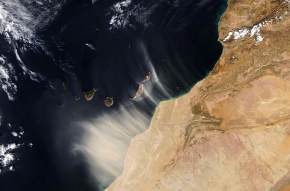 Aerial view showing a wash of pale dust sweeping from a brown landmass out over the dark ocean