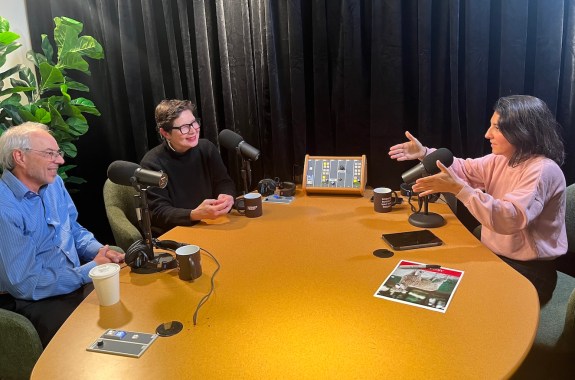 A woman and a man sit on the left side of a table, talking to a woman on the right, against a black backdrop