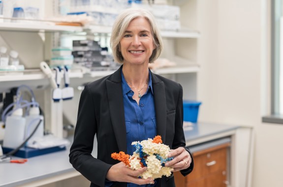 A woman in a black blazer stands in a lab smiling, holding a molecule model