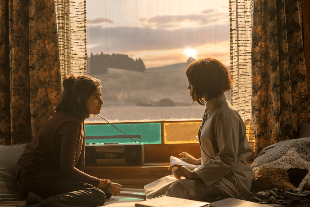 two girls sit on the floor facing each other, in front of a window with the sun setting behind them.