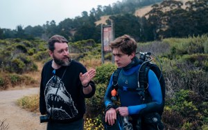 A man in a black shark sweatshirt gestures and talks to a younger man in a blue wet suit and scuba gear, standing in a shrubby outdoor area.