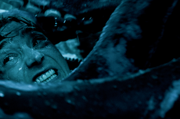 Dark close-up of a man's face as he strains and grits his teeth while being squeezed among folds.
