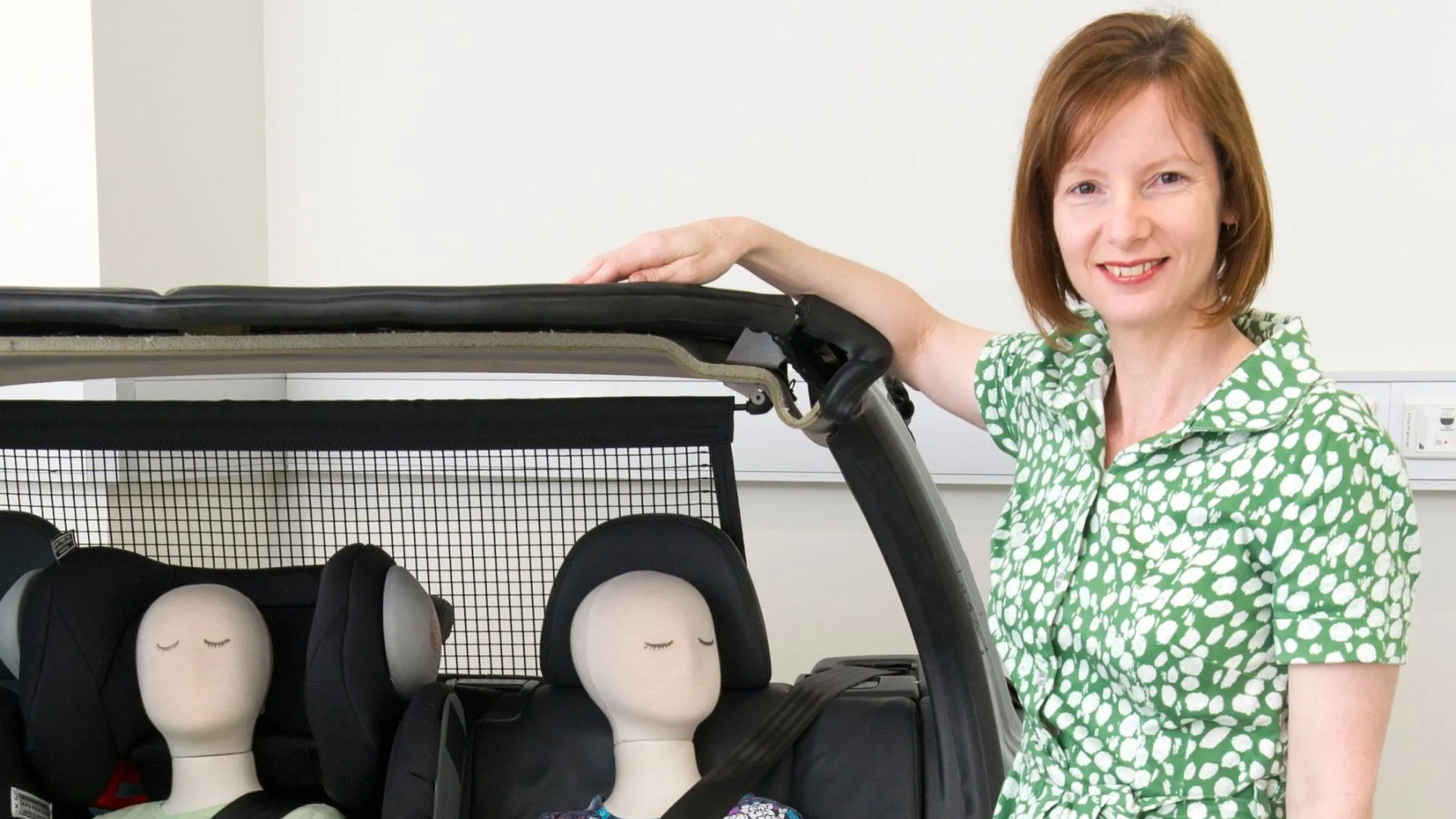 A woman in a green polka-dot dress stands beside a section of a car, resting her hand on the vehicle’s frame. Two child-sized crash-test mannequins are buckled in seats in the car.